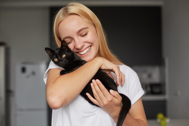 mulher loira de camiseta branca abaraçando um gatinho preto enquanto sorri.